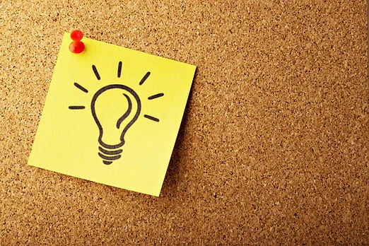 Yellow sticky note pinned to a corkboard, showing a black lightbulb icon with rays, suggesting a new idea.
