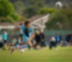 Young soccer player in light blue sprints on a grassy field as the ball flies overhead, with blurred spectators and houses behind.
