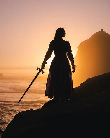 Silhouetted woman in a dress holds a sword on rocky shore at sunset, with waves and an orange sky behind her.