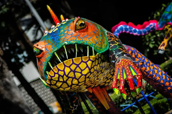 Colorful dragon sculpture with open jaws and clawed paw, painted in bright mosaic patterns against a dark outdoor background.