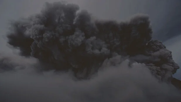 Massive dark volcanic ash cloud billows over a cloudy sky, looming ominously above a mountain.