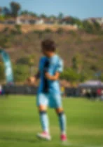 Soccer player in light blue jersey celebrates on a sunny field, with hills and houses in the background.
