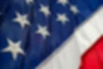 Close-up of an American flag, showing white stars on blue fabric and a red-and-white stripe, with textured stitching.