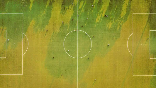 Aerial view of a soccer field with scattered players, long shadows, and green-yellow grass around the center circle and goals.
