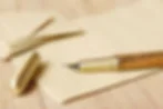 Fountain pen with wooden barrel and gold cap resting on cream lined paper, with envelopes in warm light