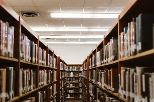 Quiet library aisle lined with tall wooden bookshelves and books under bright fluorescent ceiling lights, empty and orderly.