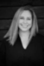 Black-and-white portrait of a smiling woman with straight hair in a dark blouse against a dark wooden wall.